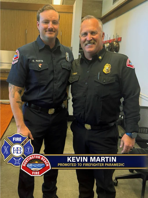 Firefighter Paramedic Kevin Martin stands next to Fire Chief Eric McCoy inside a Huntington Beach Fire Department station. Both are smiling and wearing navy blue duty uniforms. A promotional banner across the bottom of the image reads, “Kevin Martin Promoted to Firefighter Paramedic,” alongside Huntington Beach Fire Department logos. The photo was taken in a station meeting room with wood cabinetry, tables, and office chairs visible in the background.