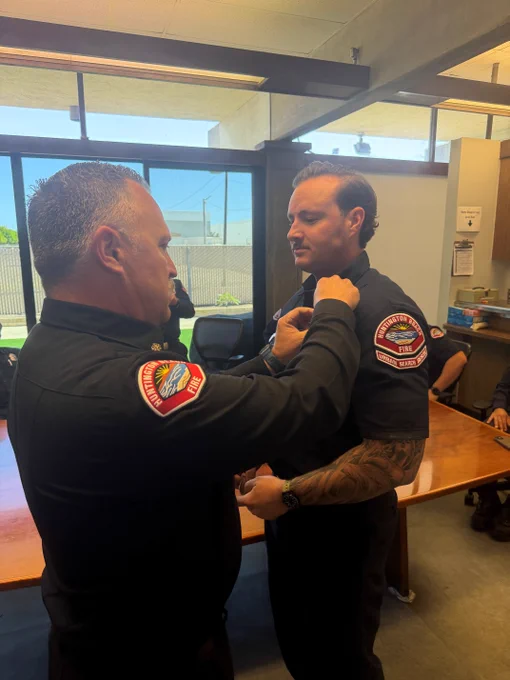 Fire Chief Eric McCoy pins Kevin Martin's new Firefighter Paramedic badge during a promotion ceremony inside a Huntington Beach Fire Department station. Chief McCoy is seen from behind while Martin stands facing him in uniform. Other department personnel are seated and standing in the background near a conference table. Large windows provide natural light into the room as the badge pinning takes place.