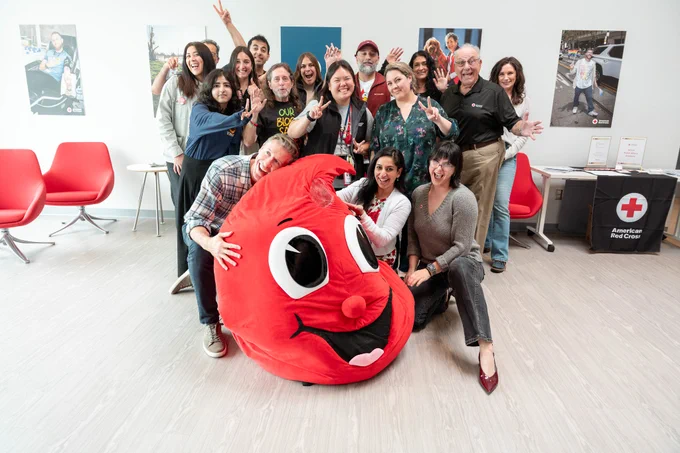 A group of people smiling and posing around a large, red blood drop mascot with a face, in a room with American Red Cross signage. Red chairs and posters are visible in the background.