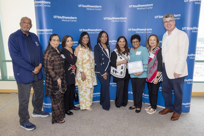 A group of people standing together, smiling in front of a UT Southwestern Medical Center backdrop. One person holds a framed certificate. The group is diverse, wearing business casual attire.