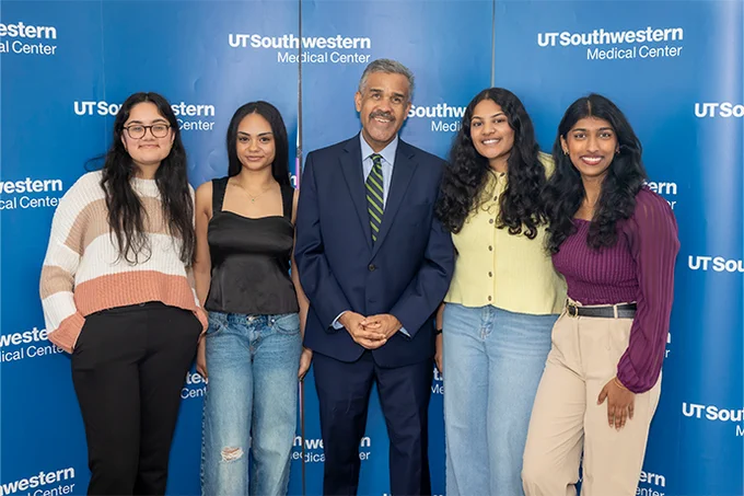 A group of five people standing in front of a blue backdrop with "UT Southwestern Medical Center" repeated on it. Four are casually dressed, and one in the middle is in a suit. They are all smiling at the camera.