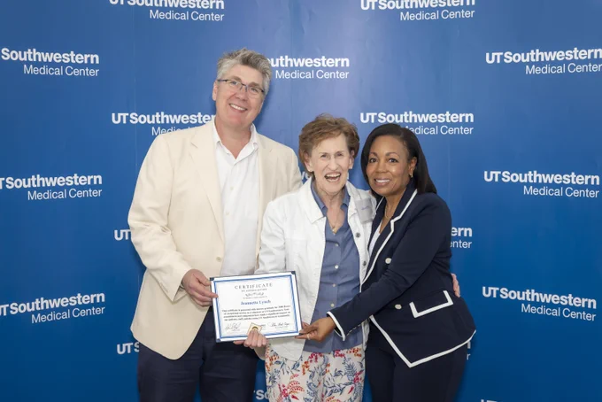 Three people smiling and standing in front of a UT Southwestern Medical Center backdrop. The person in the middle is holding a certificate.