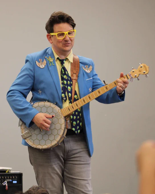 Person wearing yellow glasses, a blue blazer decorated with pins and a patterned tie playing a banjo.