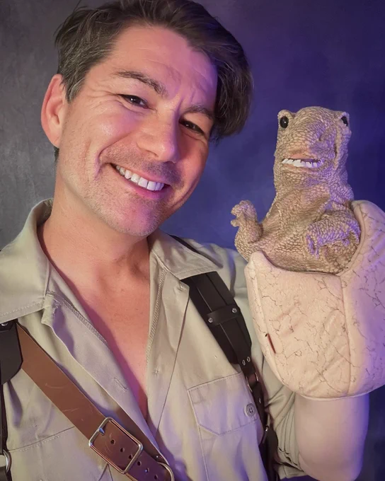 Person dressed in explorer-style shirt and leather straps holding a puppet resembling a small dinosaur coming out of a shell.