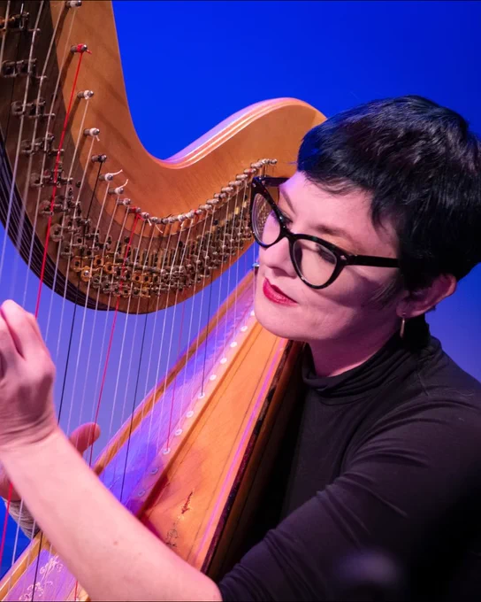 Musician plucking strings on a large wooden harp against a blue background in a close-up shot.