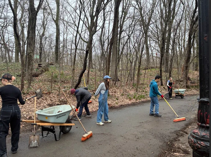 Volunteers cleaning a Prospect Park path with brooms and shovels among leafless trees on a cloudy day.