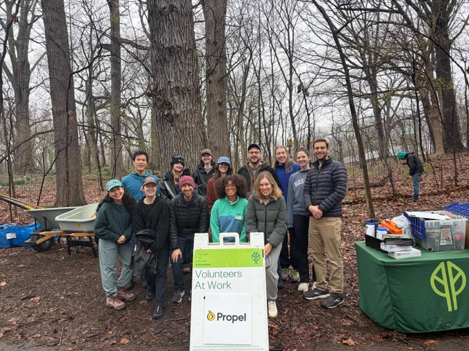 Group of volunteers posing in an early spring bare forest clearing with a sign reading 'Volunteers At Work' and a table with supplies nearby.