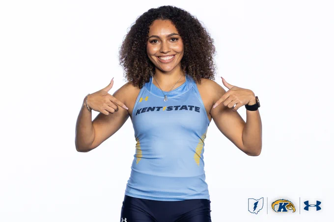 A person smiling and pointing at their "Kent State" athletic tank top. The outfit is light blue with dark shorts. Logos for Kent State and Under Armour are at the bottom.