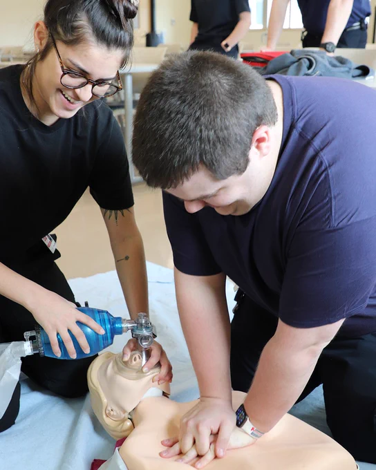 Two individuals are performing CPR on a medical training mannequin. One person is compressing the chest while the other is using a bag valve mask. Both appear focused and engaged in the training exercise.