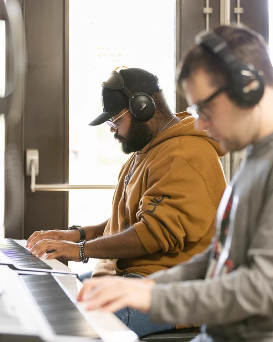 Two people wearing headphones play keyboards in a bright room. One person is wearing a cap and hoodie, while the other is wearing glasses and a sweatshirt.