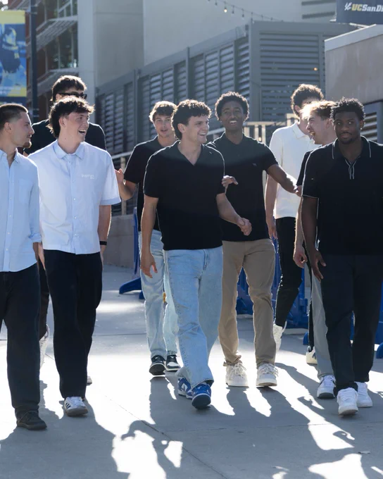 A group of people walking together outside in a sunny area near blue barricades and a building. They are casually dressed, and one person is holding a basketball. There is a "UC Santa Diego" sign visible in the background.
