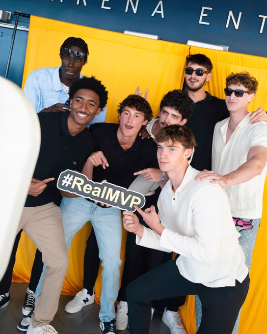 A group of people smiling and posing in front of a yellow backdrop at an event entrance. One person holds a sign that reads "#RealMVPR."
