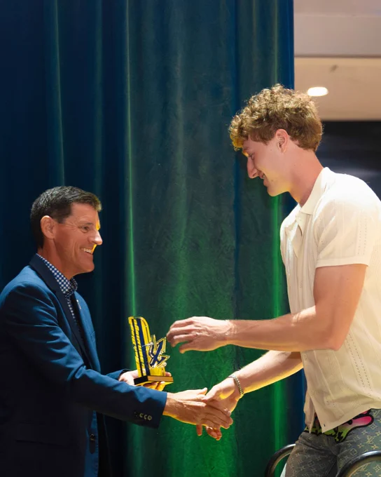 Two individuals stand on a stage exchanging a brightly colored, abstract trophy. One person is dressed in a suit, and the other in a casual shirt and patterned pants. The background features a dark curtain and part of a brightly lit ceiling.
