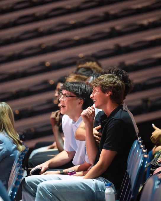 A group of people sitting in a large auditorium, engaged and smiling. The seating is tiered, and the audience appears attentive to an unseen event.