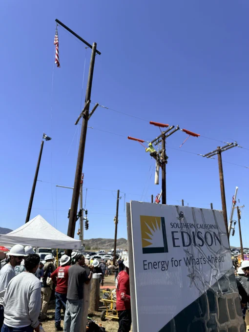 A group of people wearing safety helmets gather near utility poles under a clear blue sky. A person is working on one of the poles. A Southern California Edison sign is in the foreground. An American flag is visible at the top of a pole.