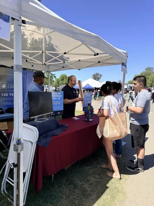 A group of people stand at a booth under a white canopy at an outdoor event. The booth is operated by Southern California Edison and features a sign about its 140-year anniversary and a colorful spin wheel on the table. In the background, more attendees can be seen.