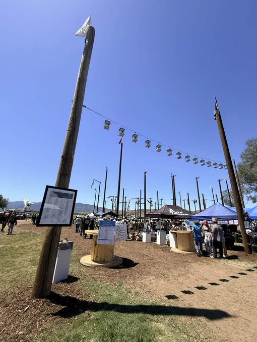 An outdoor event with tall wooden poles used for competitive climbing activities. Many people are gathered around tents and tables. Decorative flags are strung between poles. The setting is under a clear blue sky with mountains in the background.
