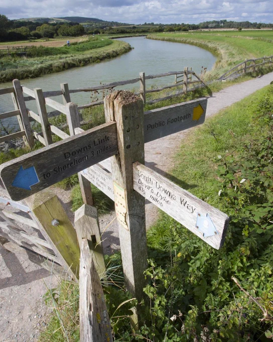 Wooden fingerpost pointing to the Downs Link and South Downs Way, with green countryside and the River Adur in the background.