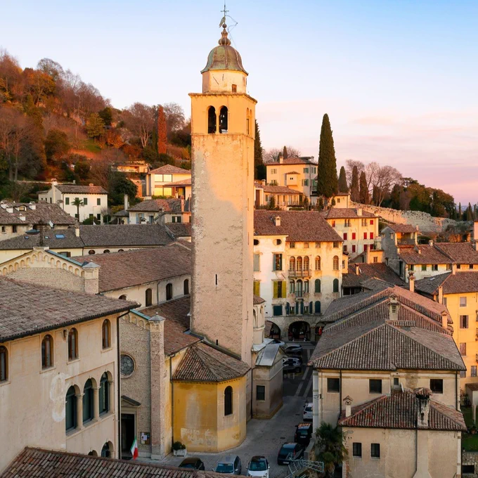 A scenic view of a historic Italian town at sunset, featuring a tall bell tower surrounded by traditional buildings with terracotta roofs. The landscape includes hillside trees, and the sky has a pink hue.
