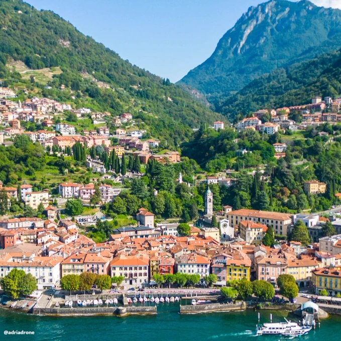 Aerial view of the colorful town of Bellano on the shores of Lake Como in Italy. The town features vibrant buildings, a waterfront promenade, and lush greenery. Mountains rise majestically in the background under a clear blue sky. A boat is docked at the lake.