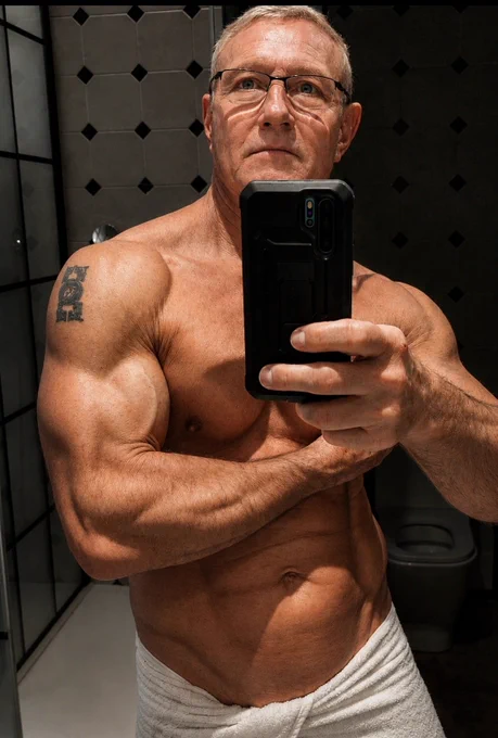 Mirror selfie of Pascal Saint James, a 63-year-old man showing a muscular natural physique after training, with defined arms and shoulders, wearing a towel in a bathroom. Fitness after 60, healthy aging, longevity, discipline and lifestyle