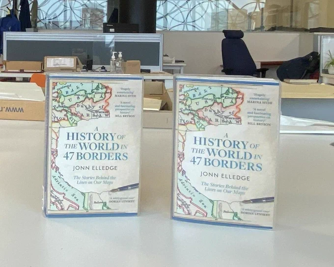 Two copies of a book on a table with front covers facing on. Title of the book is A History of the World in 47 Borders by Jonn Elledge. Outline of a historic map on the far left of the page. An office space behind the table on which the books are standing. Reflection of ceiling lights and a window with a metal filigree casing in the far background.