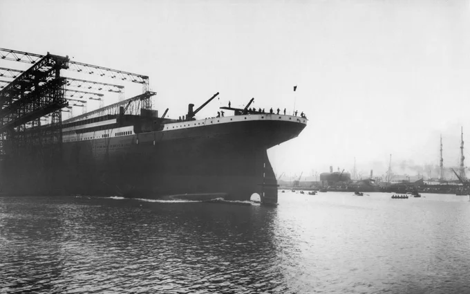 A large ship, the RMS Titanic, is seen partially constructed in a shipyard. The ship is in the water with scaffolding above, surrounded by a few small boats. The background shows other ships and industrial structures under a cloudy sky.