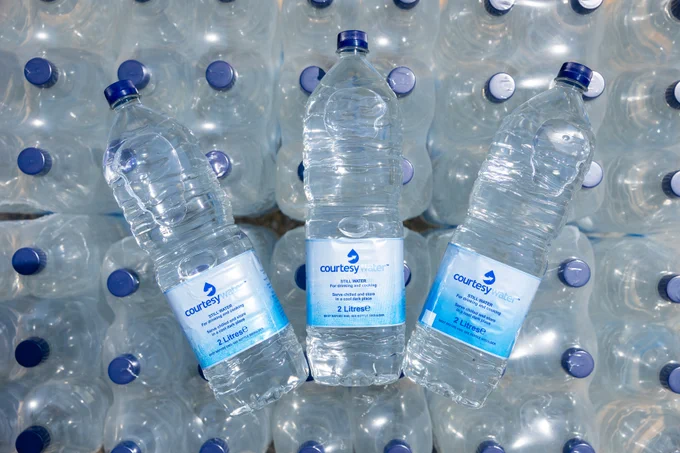A close-up of three plastic water bottles labelled  "Courtesy Water," angled towards the camera. They are set against a blurred background of many stacked, clear plastic water bottles with blue caps.
