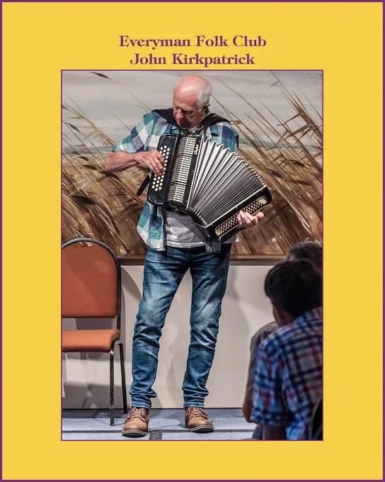 Photo of John Kirkpatrick, standing playing button accordion. John is wearing a white t-shirt beneath a shirt with a pattern made up of large blue squares, and blue jeans.