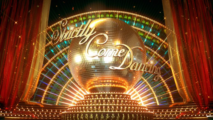 A glittering ballroom stage with a large, illuminated disco ball featuring the "Strictly Come Dancing" logo. The background is adorned with colorful lights and shimmering decorations.