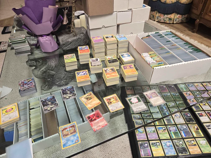 Photo of a large Pokémon card sorting and organization project spread across a glass coffee table. Multiple stacks of Pokémon cards are arranged by set, type, or category, with several thousand cards visible in sorting trays, storage boxes, and tall card piles. White card storage boxes filled with sleeved and unsleeved cards sit on the right side of the table, many separated by labeled dividers.

Several featured cards are laid out individually across the workspace, including vintage-era Pokémon cards and modern full-art cards. Open binders in the lower right display pages filled with holographic and reverse-holographic Pokémon cards in protective sleeves. Additional long boxes filled with cards are visible in the foreground.

In the background are more storage boxes waiting to be sorted, stacks of trading cards, and household furnishings. The image conveys an extensive card cataloging effort involving thousands of Pokémon cards being sorted, organized, and prepared for binders.