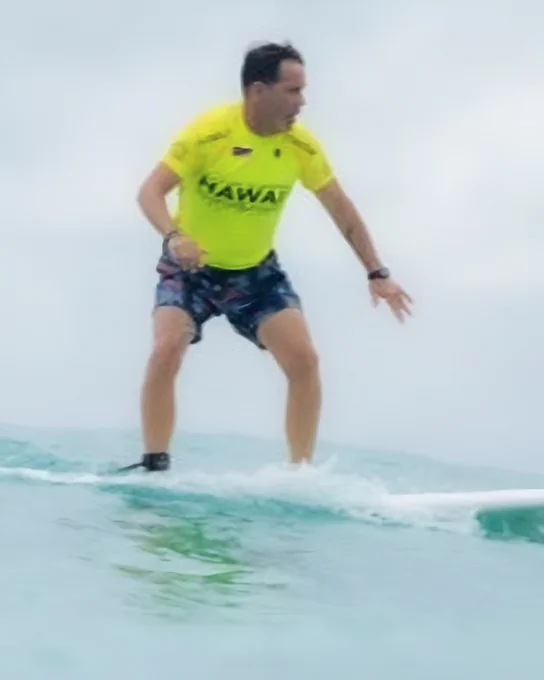 A person in a bright yellow shirt surfs on a small wave, balancing on a surfboard in the ocean.