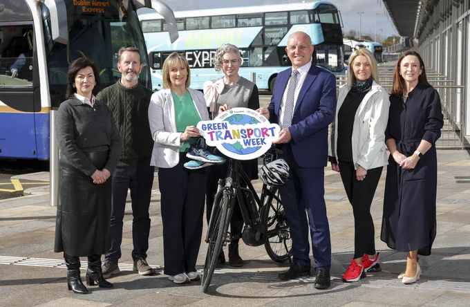 Seven smiling people, 4 women and 3 men, stand by a bicycle and blue running shoes. They hold a cloud-shaped sign with a globe and 'GREEN TRANSPORT WEEK'.