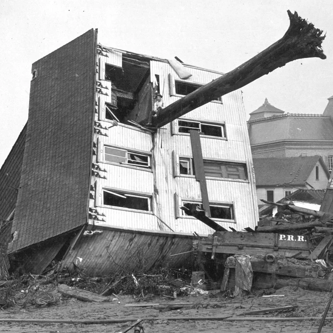 A black and white photo of a house turned on its side after the South Fork Dam failed and unleashed a massive flood. A large tree sticks out of the house with a boy sitting on it. 