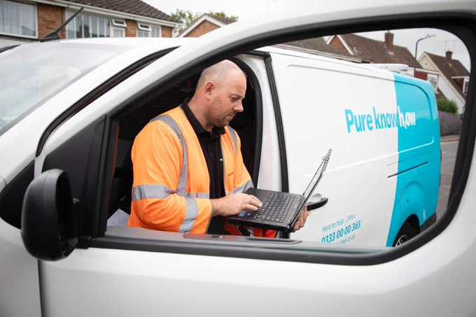 Photograph of a South East Water employee wearing an orange hi-vis, standing next to an open door of a South East Water van using his laptop.