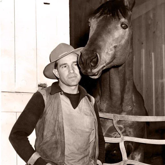 George Woolf stands beside Seabiscuit at a stable doorway. Woolf wears a wide-brimmed hat, jacket, shirt, and trousers, with one hand on his hip. Seabiscuit stands behind a rope barrier with his head lowered near Woolf’s shoulder. The black-and-white photo shows wooden stable walls and straw on the floor.
