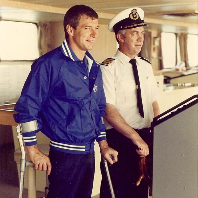 Image shows Rick Hansen onboard the vessel with a captain during the Man in Motion tour. Rick is standing with arm braces, wearing a blue jacket and jeans. The captain to his right is wearing a white captains hat, a white short sleeve button up, and a black tie. Both are looking into the distance.