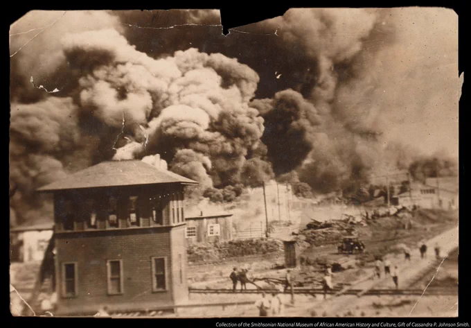 A black-and-white photograph of the Greenwood District of Tulsa, Oklahoma burning during the Tulsa Race Massacre in 1921. The image depicts a building, several people, and large plumes of smoke rising and filling the air. The photograph is bent and weathered at the edges. An image credit is in the bottom right.