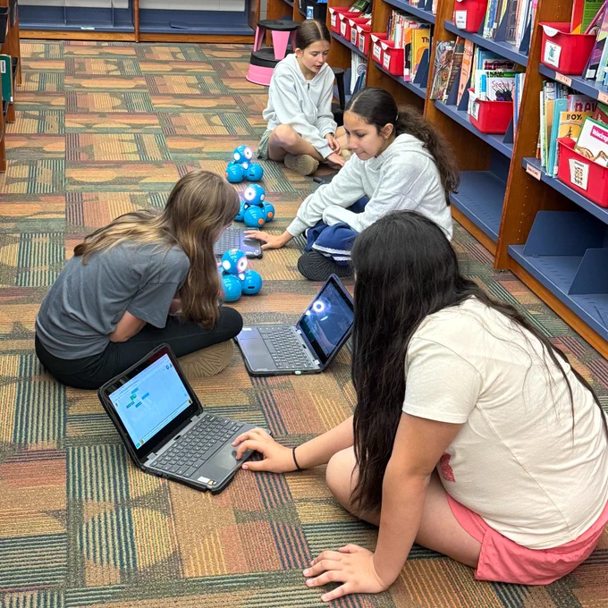 Four students sitting on a library floor working on laptops and programming blue robots lined up in front of them.