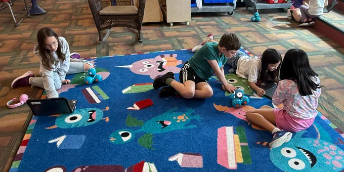 Four children sitting and lying on a colorful classroom rug, engaged with small robots and a laptop during a hands-on learning activity.