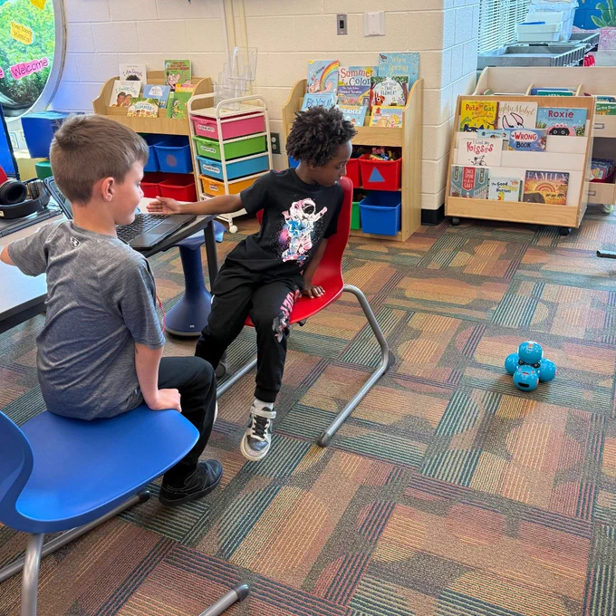 Two children seated in a colorful classroom with bookshelves and a small blue robot on the carpet nearby.