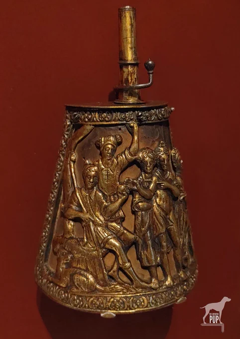 Late 16th-century Italian powder flask  made of gilt brass over fabric-covered wood depicting Paris of Troy with a dog, an attendant, and three Greek goddesses. 