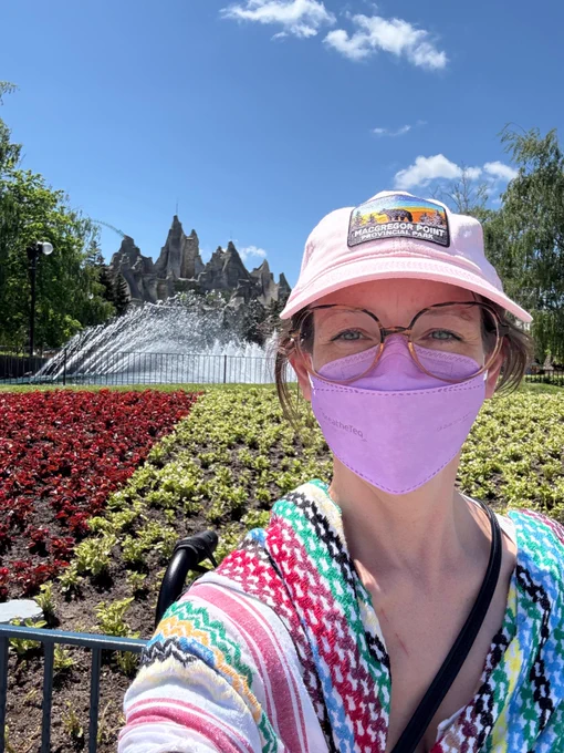 Photo of me wearing my rainbow hirbawi keffiyeh, a pink macgregor point provincial park hat and a lavendar breatheteq kn95 respirator mask while in front of the Canada’s Wonderland mountain and main fountain