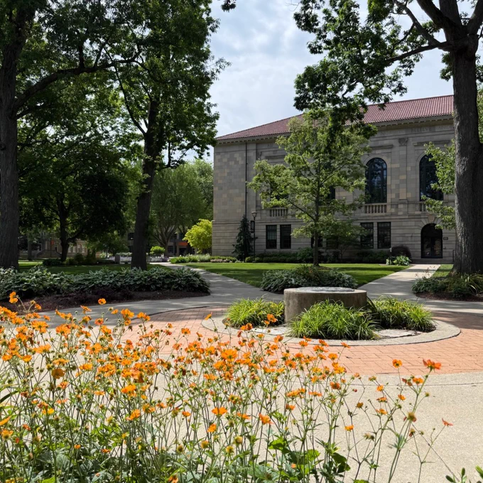 A scenic view of a campus area at Bowling Green State University, featuring a building with arched windows in the background. Orange flowers and large trees surround the area, with paved pathways visible.