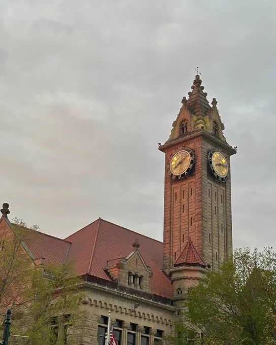 A tall, ornate clock tower with a pointed roof and illuminated clock faces stands against a cloudy sky. The building is made of stone with red-tiled roofs, and trees with green leaves are visible in the foreground.
