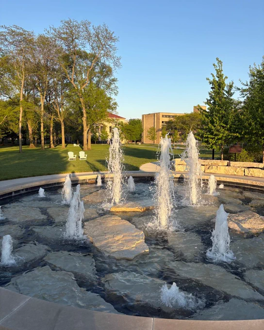 A stone fountain with multiple water jets is set in scenic parkland on the BGSU campus. In the background, there are lush green trees, part of a red-roofed building, and another tan brick building. Two white chairs are placed on the grass near the treeline beneath a clear blue sky.