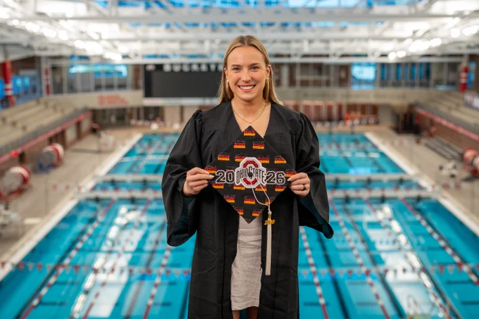 A female in graduation attire stands in an indoor swimming facility holding a decorated graduation cap featuring "2026" and Ohio State University's logo. The background shows numerous swimming lanes and seating areas.