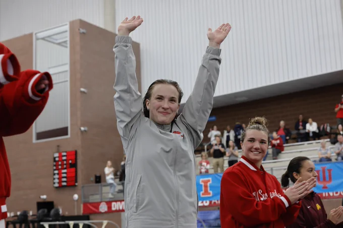A person in a gray jacket stands with arms raised, smiling at an indoor sports venue. They are on a platform with people wearing red jackets. Banners with "IU" and "Championships" are visible in the background.