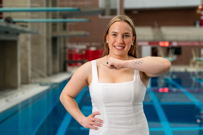 A person in a white outfit stands confidently by a swimming pool, displaying an Olympic rings tattoo on their forearm.
