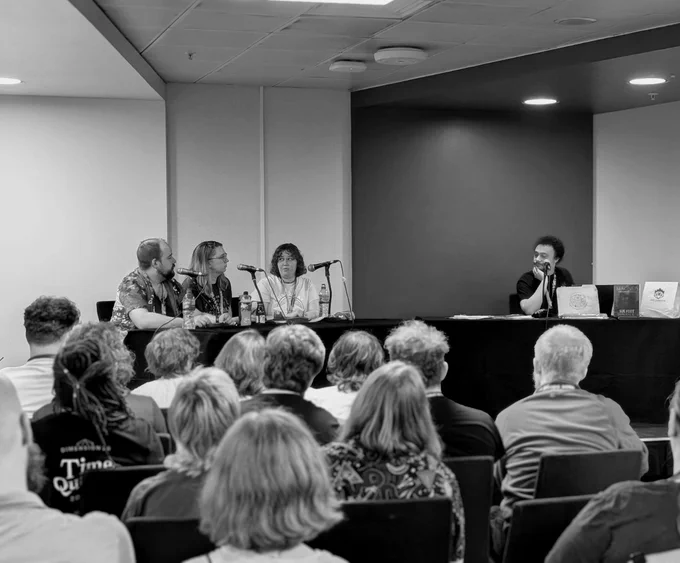 A black and white photograph of an audience seated in front of a stage. On the stage, seated at a table from left to right is Jonathan Sims, Anusia Battersby, Sydney Engelstein, and Gregor, the GM. There are microphones on the table, as well as some Magnus merchandise.